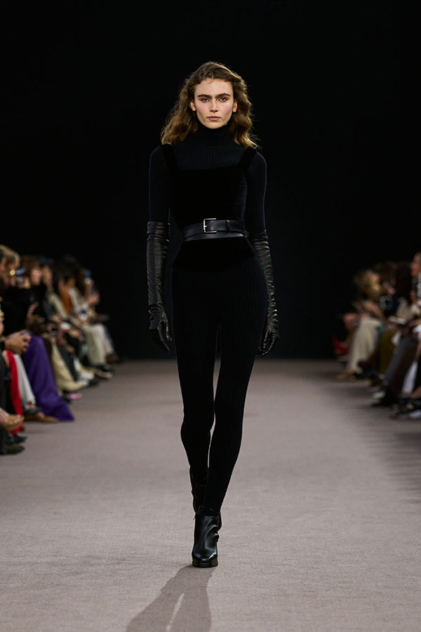 Desfile Max Mara otoño-invierno 2025 - Look negro con jersey, top bustier de terciopelo, cinturón, leggings y botines. 